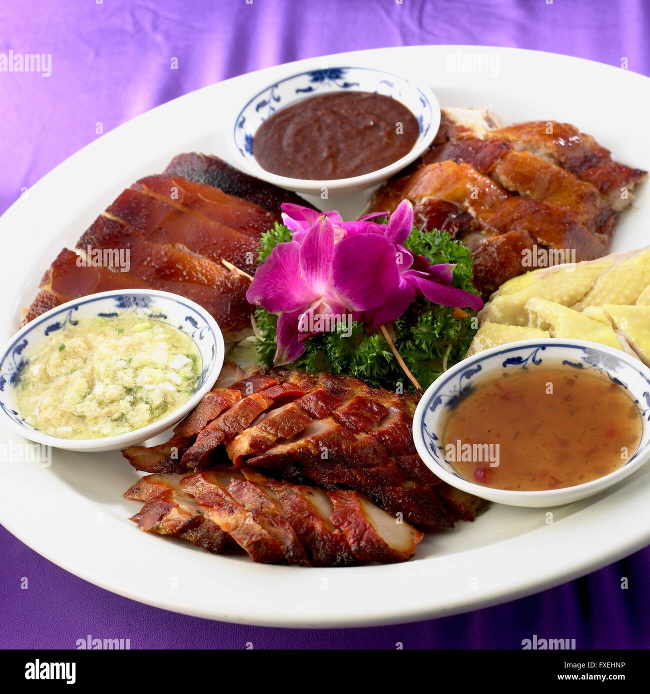 Chinese roast meats, including cuts of suckling pig, duck, pork, and chicken, served on platter with dipping sauces and garnished with parsley and flowers Stock Photo