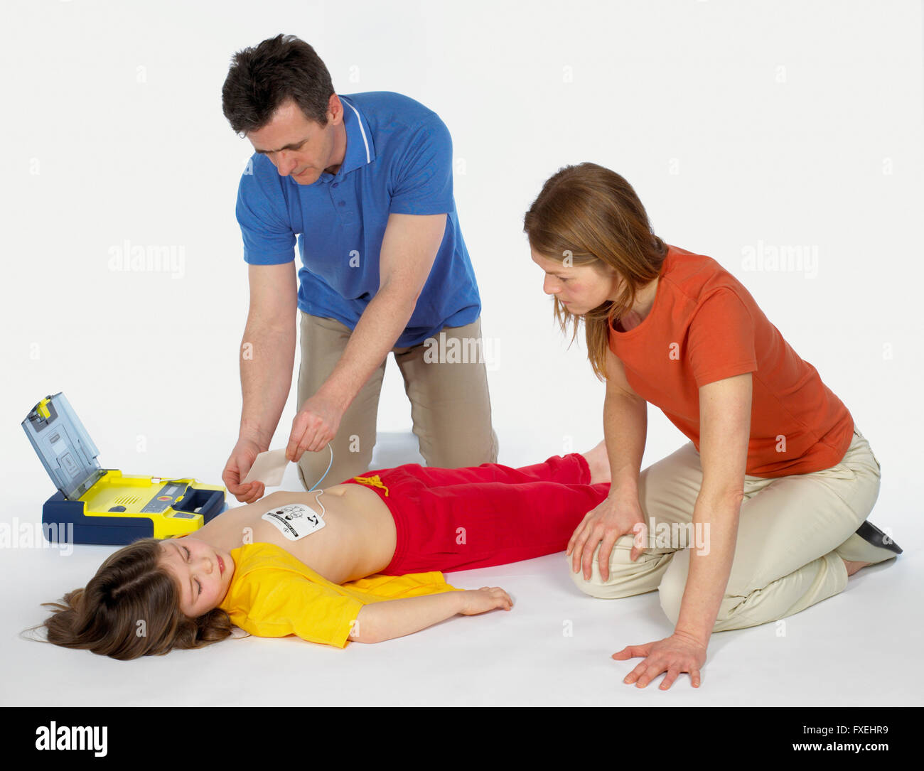 First aid, using paediatric defibrillator, elementary age girl lying on floor unconscious, mature man attaching defibrillator pads to girl's chest, mid adult woman watching Stock Photo