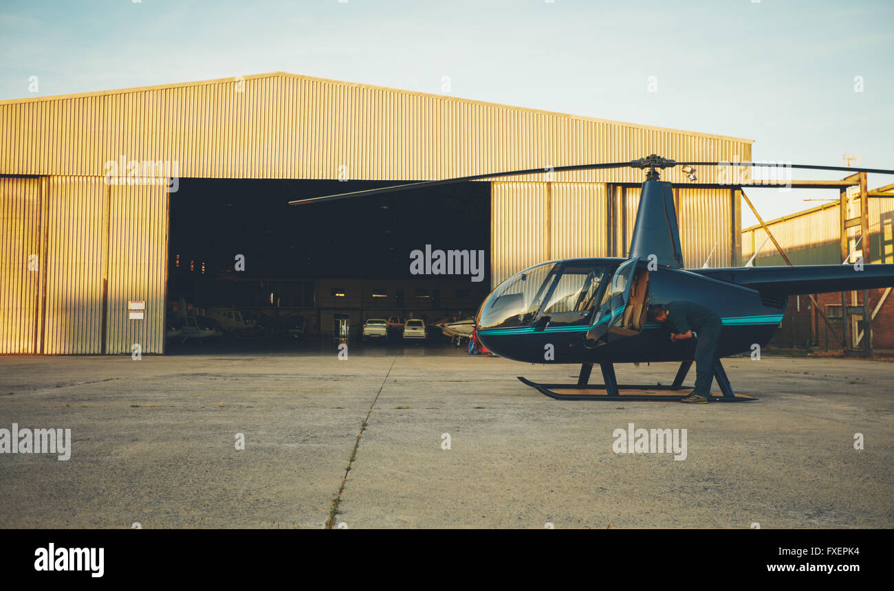 Helicopter pilot inspecting his aircraft  before take off. Helicopter is parked outside airplane hangar at airport. Stock Photo