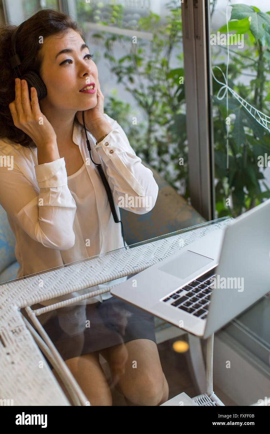 Asian woman enjoying music in headphones sitting with a laptop in a cafe. Stock Photo