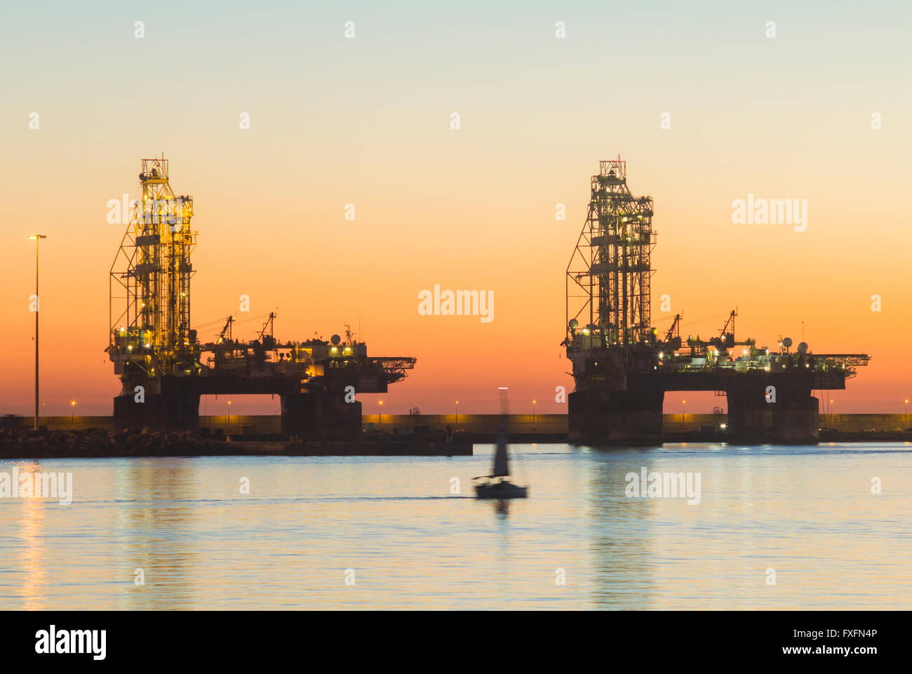 Las Palmas, Gran Canaria, Canary Islands, Spain, 15th April 2016. Weather: A yacht sails past mothballed oil rigs at sunrise in Las Palmas port on a glorious Thursday morning in Las Palmas, the capital of Gran Canaria. PICTURED: Las Palmas port is used by a number of oil companies for repairs, resupplying, and in the the case of some of the rigs/platforms in these images, for mothballing rigs for long periods due to the fall in oil prices and over supply of crude oil. Credit:  Alan Dawson News/Alamy Live News Stock Photo