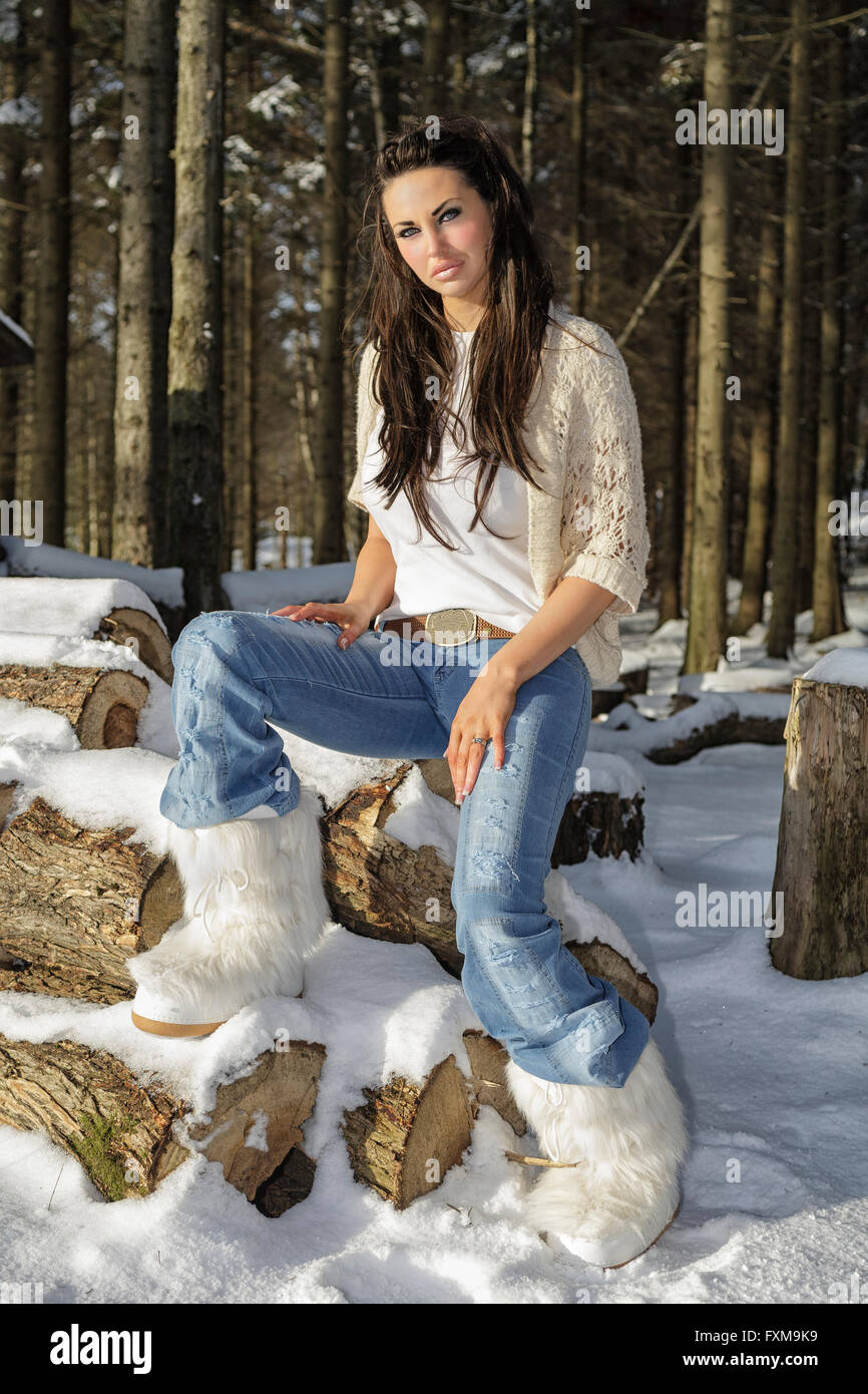 Attractive beautiful young brunette woman in fur winter snow boots sitting on wood logs in forest outdoors in winter  Model Release: Yes.  Property Release: No. Stock Photo