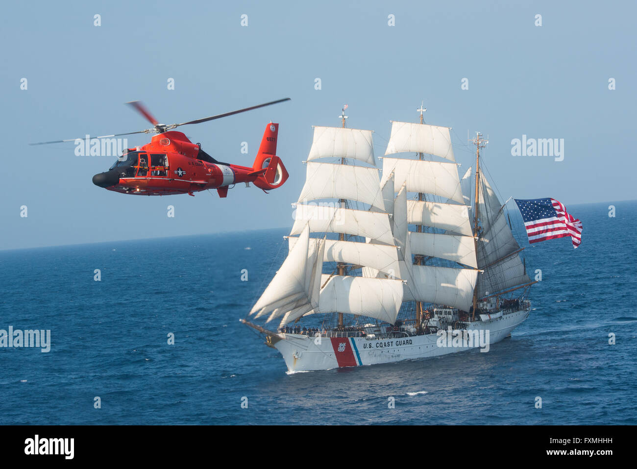 A Coast Guard MH-65 Dolphin helicopter past the Coast Guard barque Eagle with full sails July 30, 2015 in the Atlantic Ocean. The photo is a recreation of the U.S. Postal Service Forever Stamp created by William S. Phillips, an aviation artist. Stock Photo