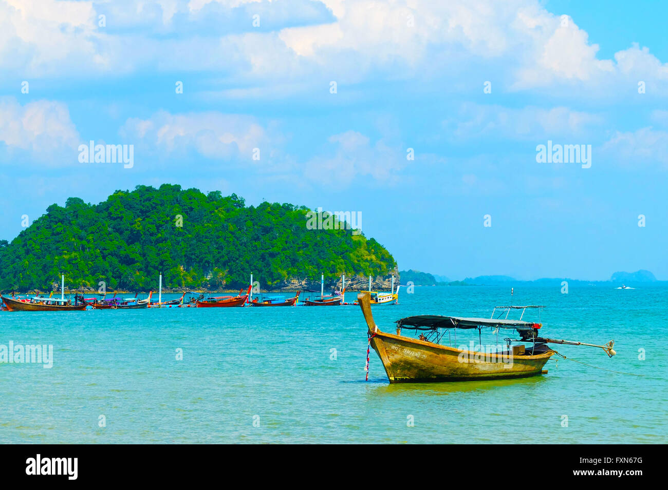 Thai long-tail boat anchored in sky-blue water with the more long-tail boats and a sea cliffs covered  green in the background. Stock Photo