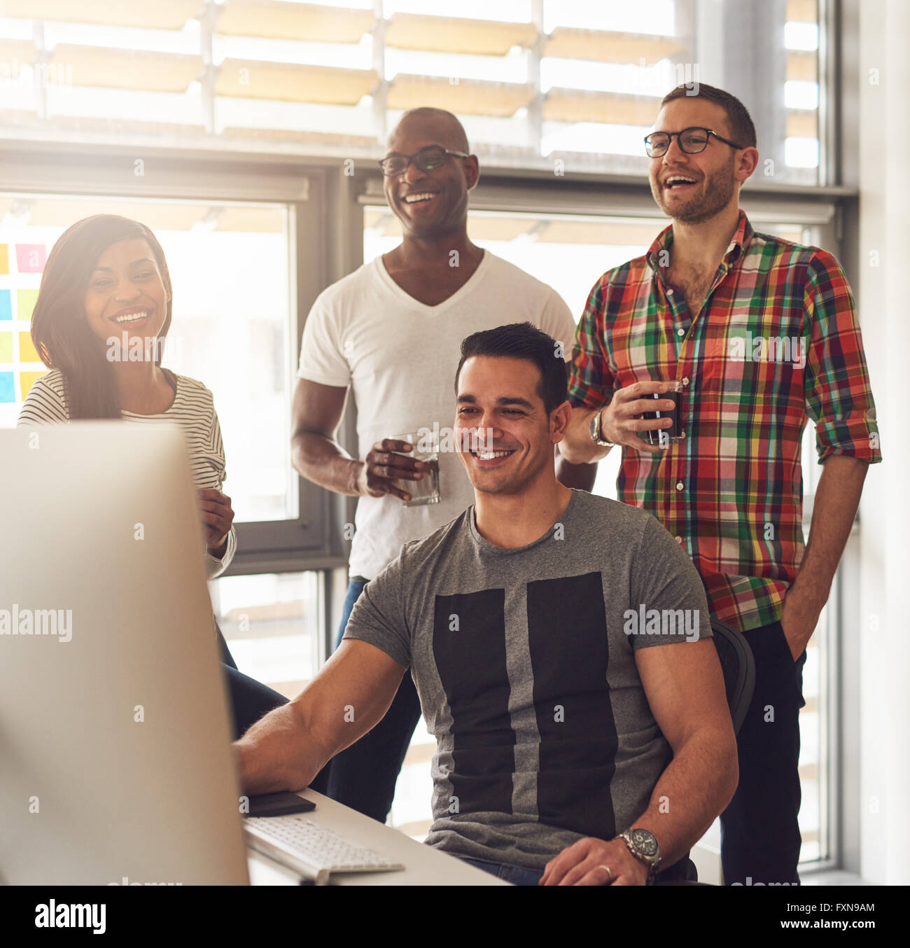Happy quartet of Black, Hispanic and Caucasian young adults at desk smiling and laughing while looking at computer monitor with Stock Photo