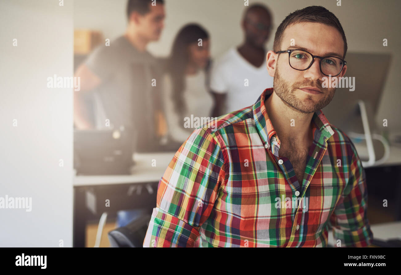 Serious single male business owner wearing multi-colored flannel button shirt with three workers on computer in background Stock Photo