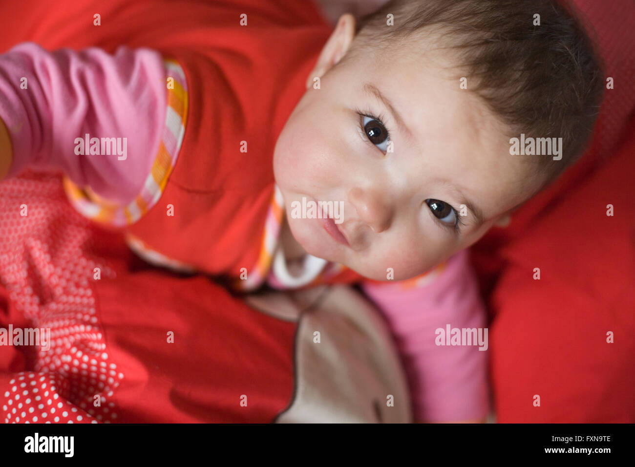 Little baby girl with beautiful dark eyes looking up just after awakening. Red bed clothes background Stock Photo