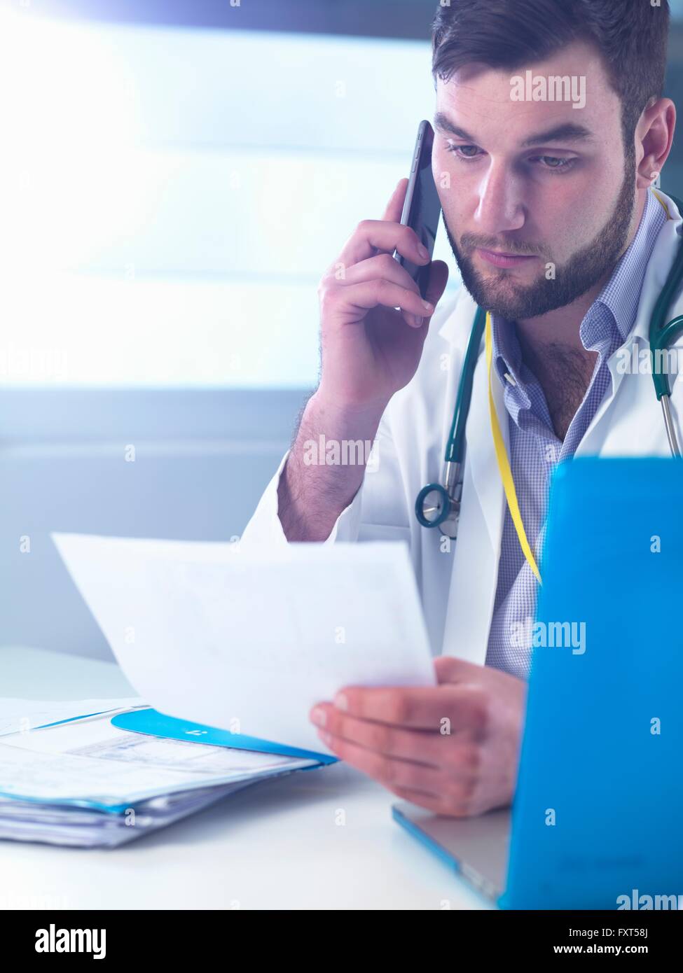 Junior doctor using smartphone whilst reading medical records in clinic Stock Photo
