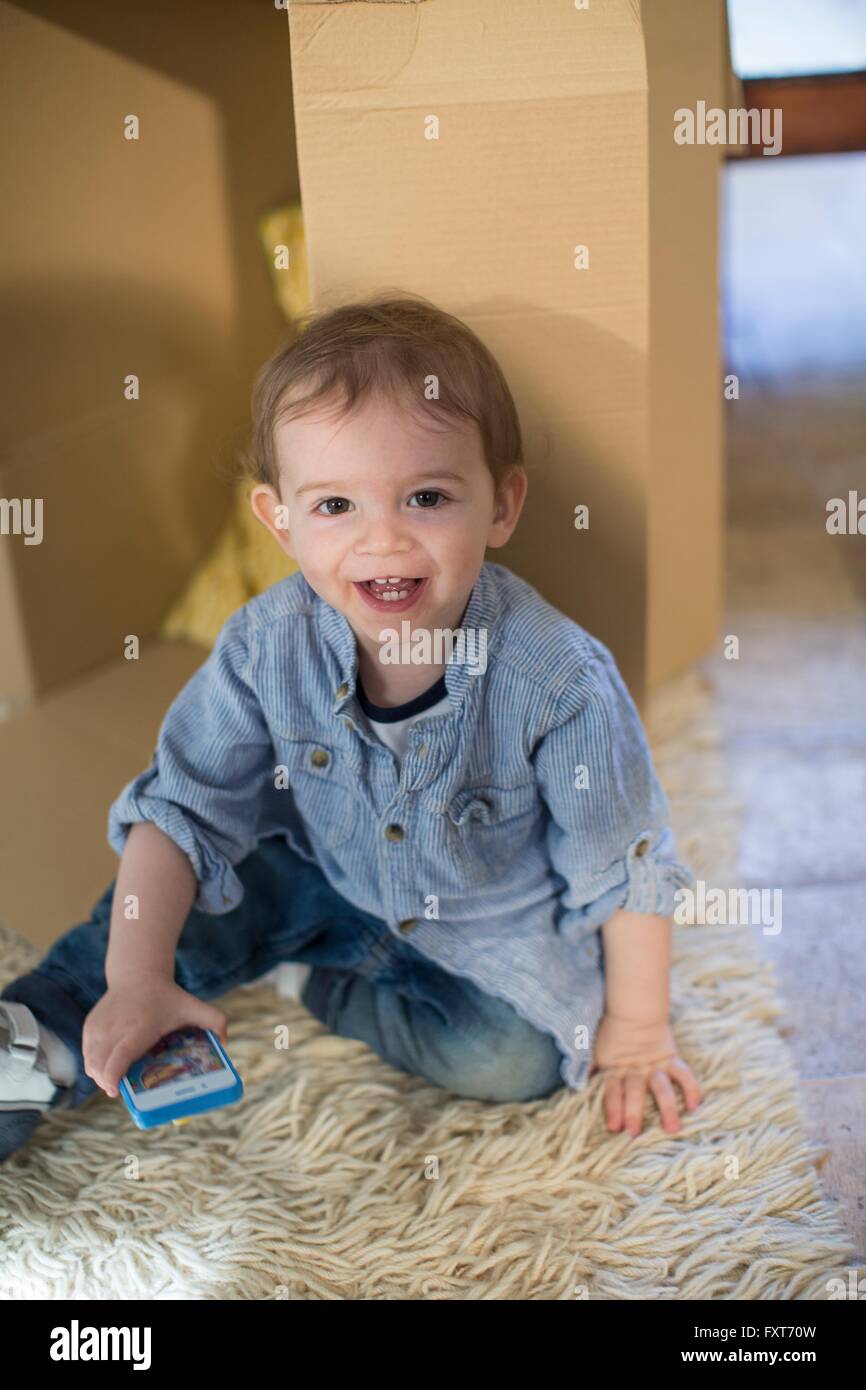 Baby boy sitting in front of cardboard box holding smartphone looking at camera smiling Stock Photo