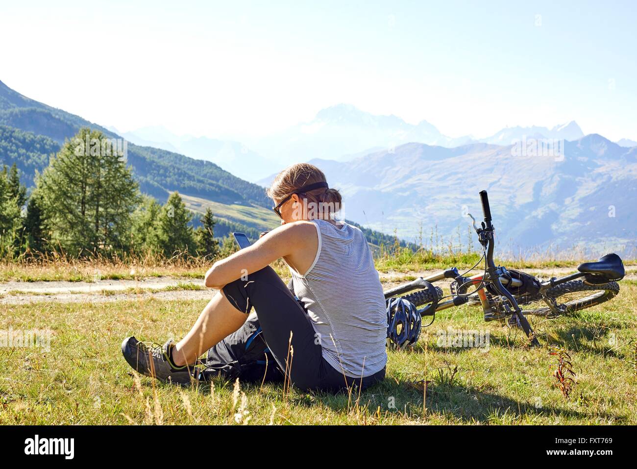 Female mountain biker sitting in mountain landscape reading smartphone texts, Aosta Valley, Aosta, Italy Stock Photo