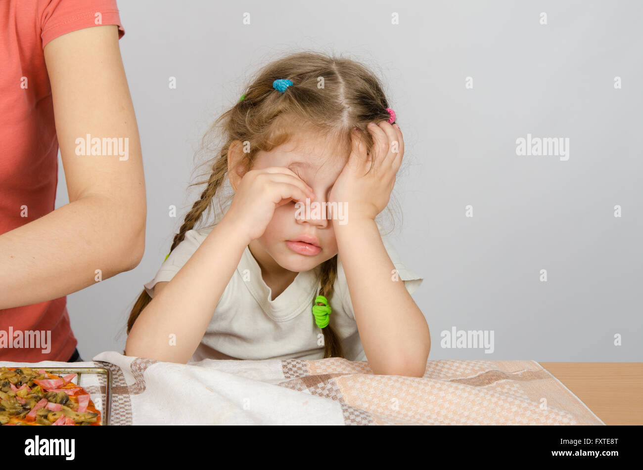Little girl wearily rubbing his eyes at the kitchen table while Mom cooks Stock Photo
