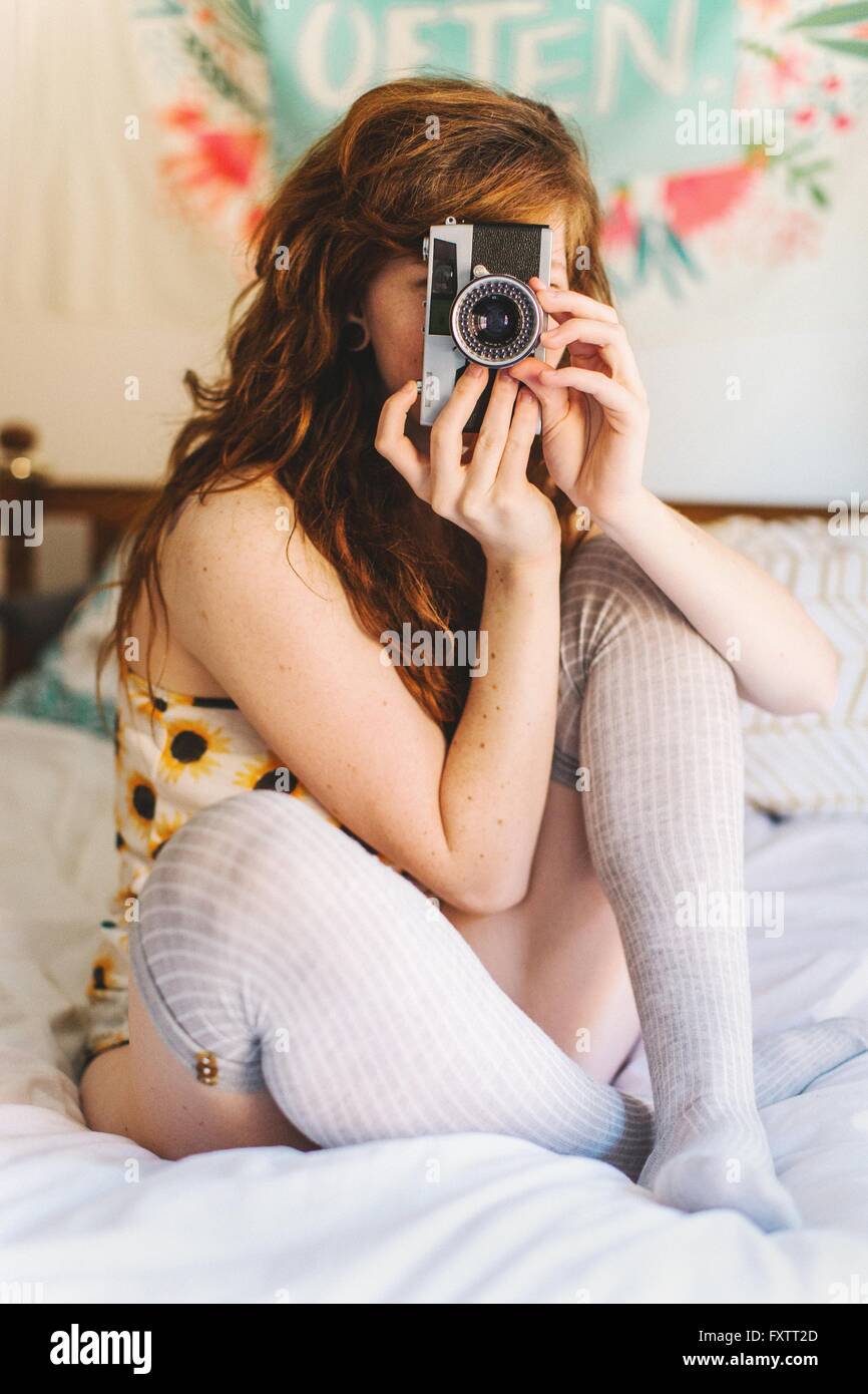 Young woman sitting on bed, looking through SLR camera Stock Photo