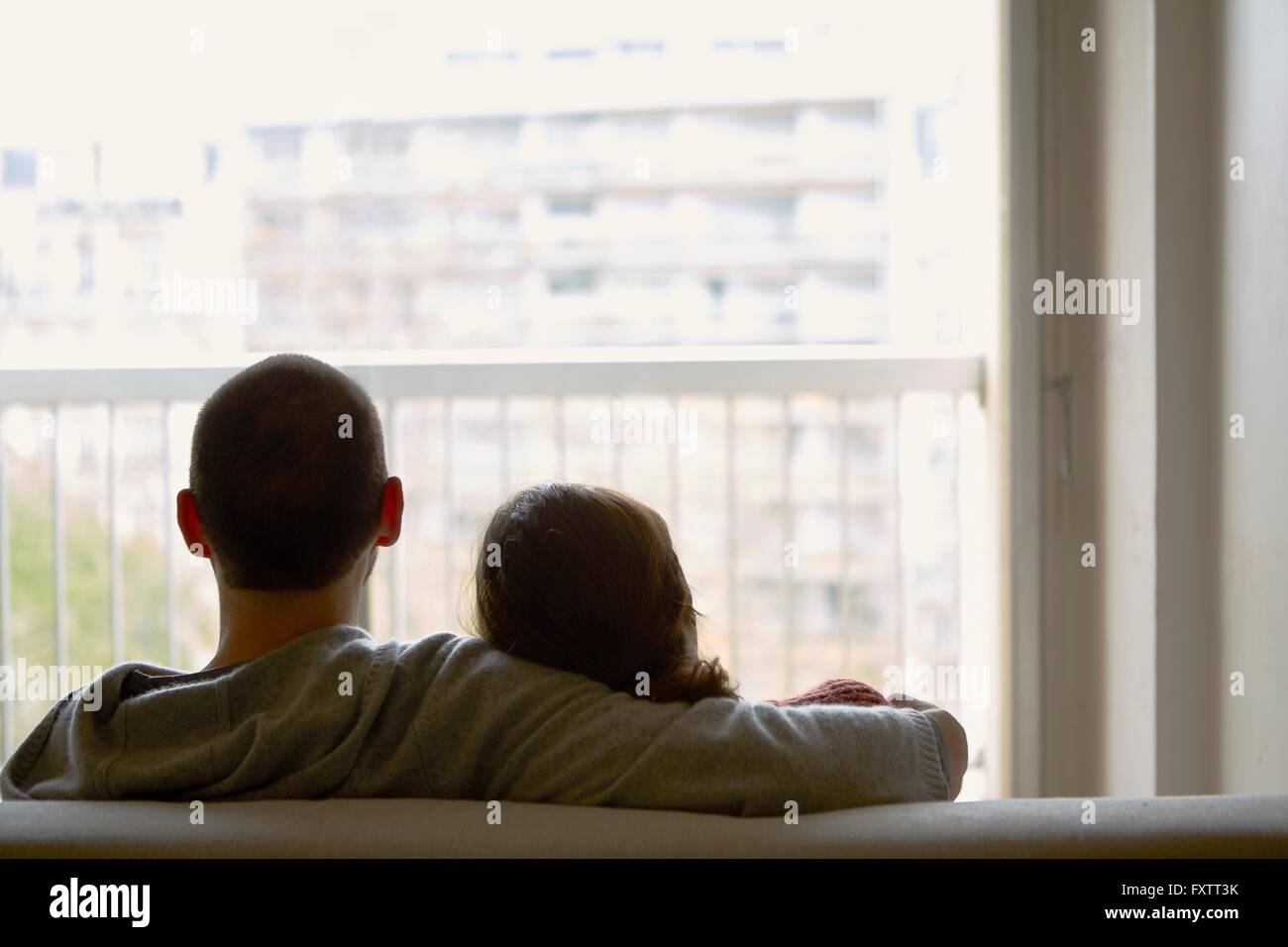 Rear view of couple sitting on sofa looking out of window at apartment building Stock Photo