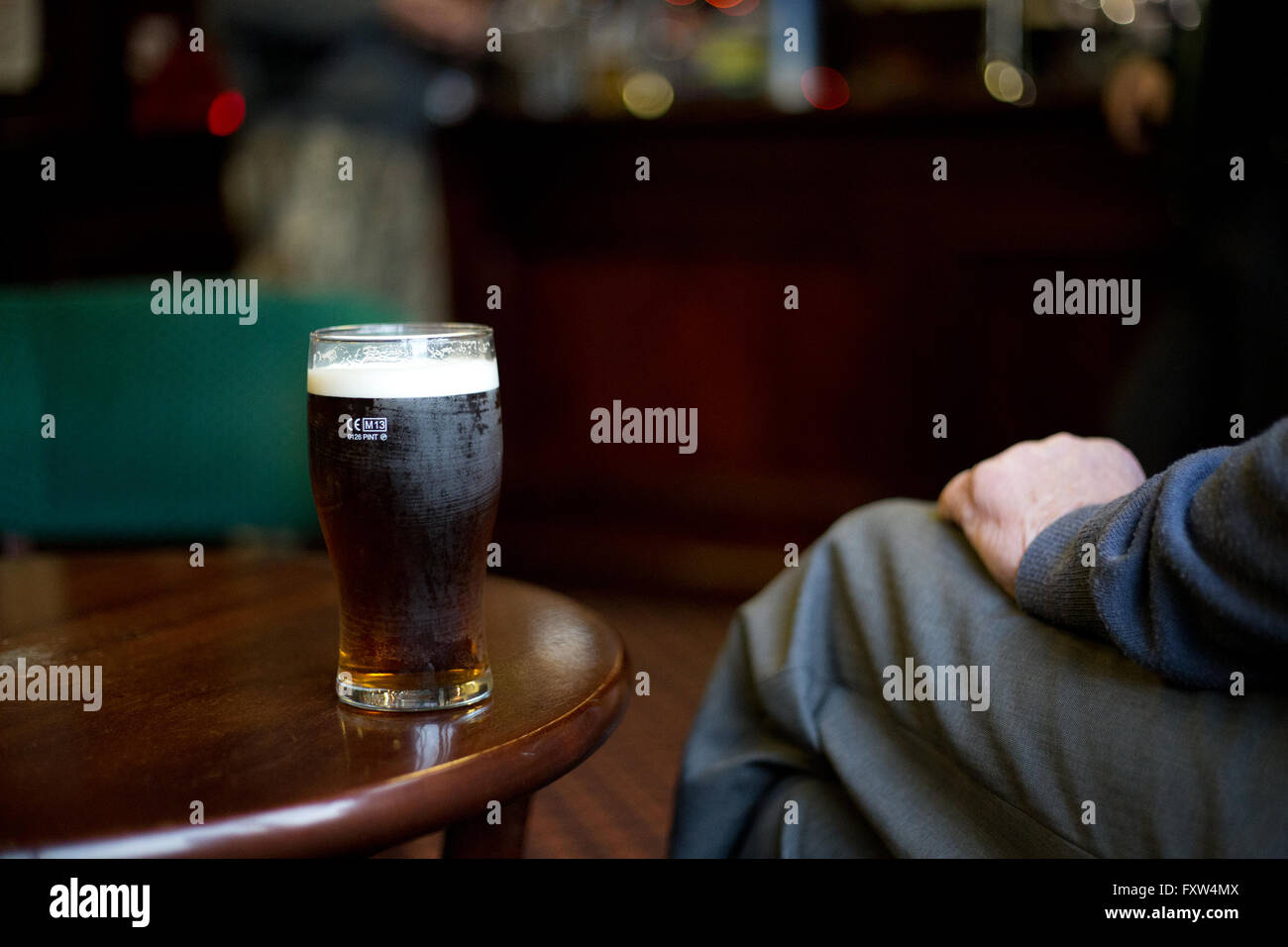 A photograph of a pint of beer on a table with a man's hand and leg in view in a pub Stock Photo