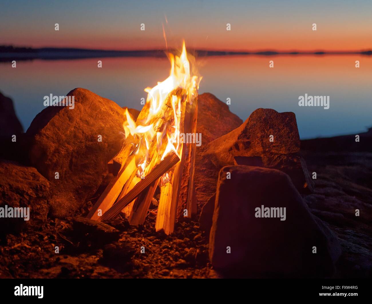 Fire and flames of atmospheric campfire on the beach at night. Still water of the lake on the backgound with warm colors of suns Stock Photo