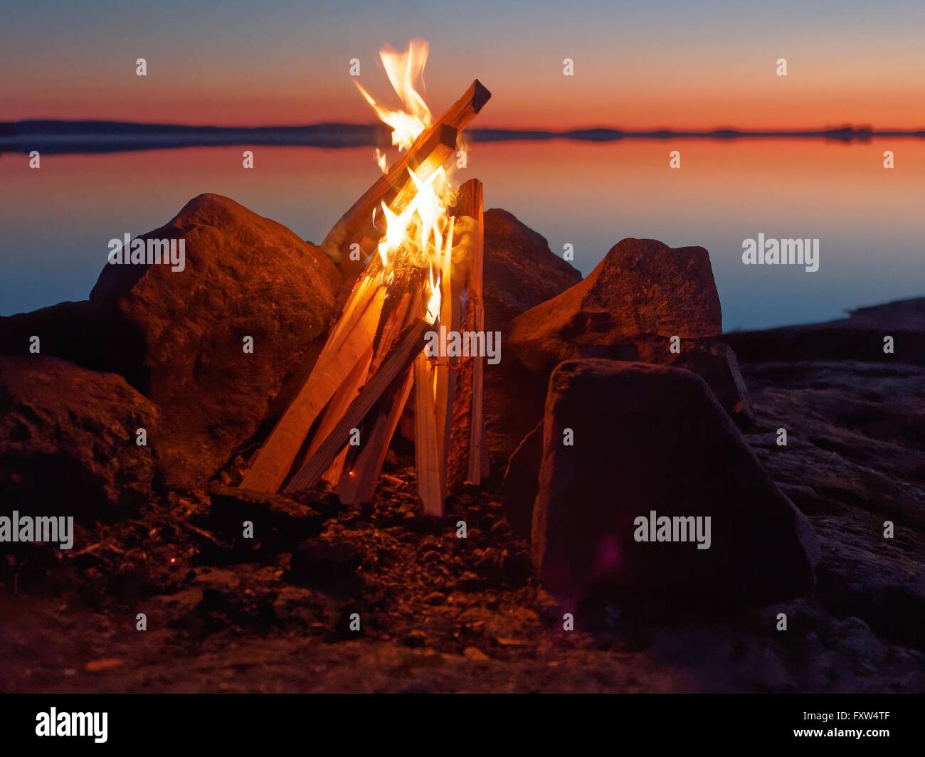 Fire and flames of atmospheric campfire on the beach at night. Still water of the lake on the backgound with warm colors of suns Stock Photo