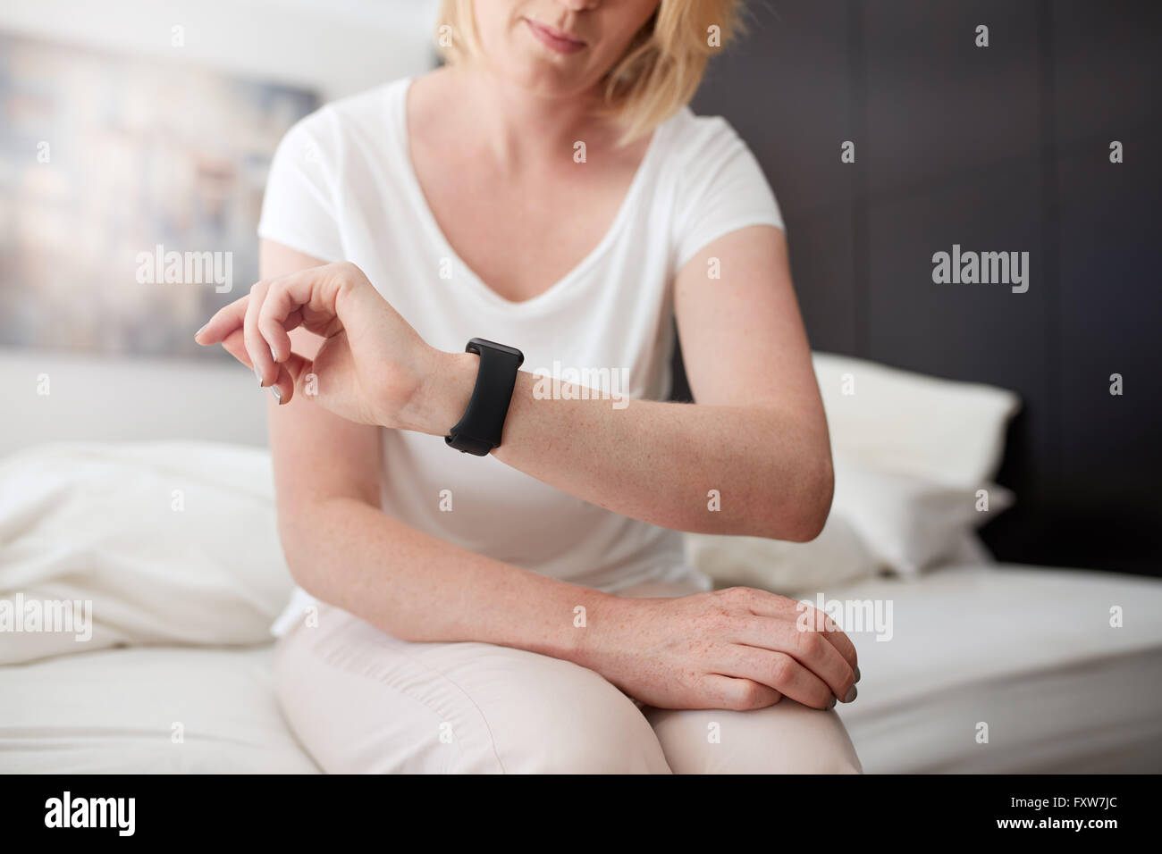 Cropped shot of a woman sitting on bed checking time on her wrist watch, she is in bedroom at home. Stock Photo