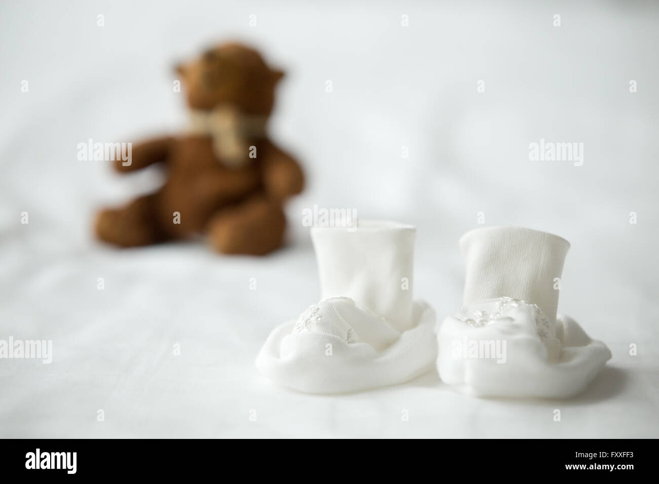Close-up of white tiny booties for newborn baby on the bed. Cute teddy bear on the background. Maternity concept Stock Photo
