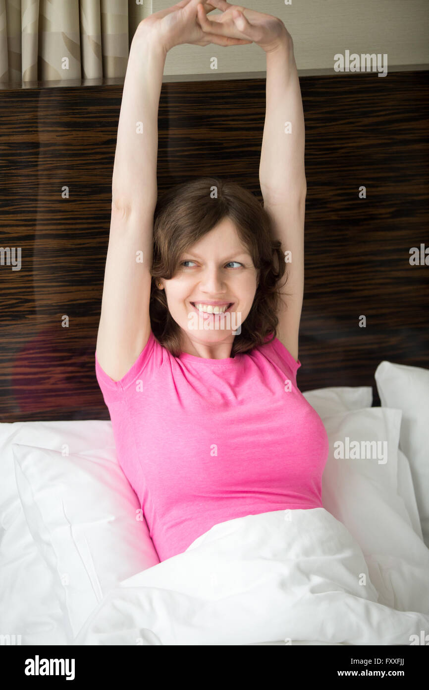 Portrait of cheerful young woman lying in bed after waking up and stretching with pleasure