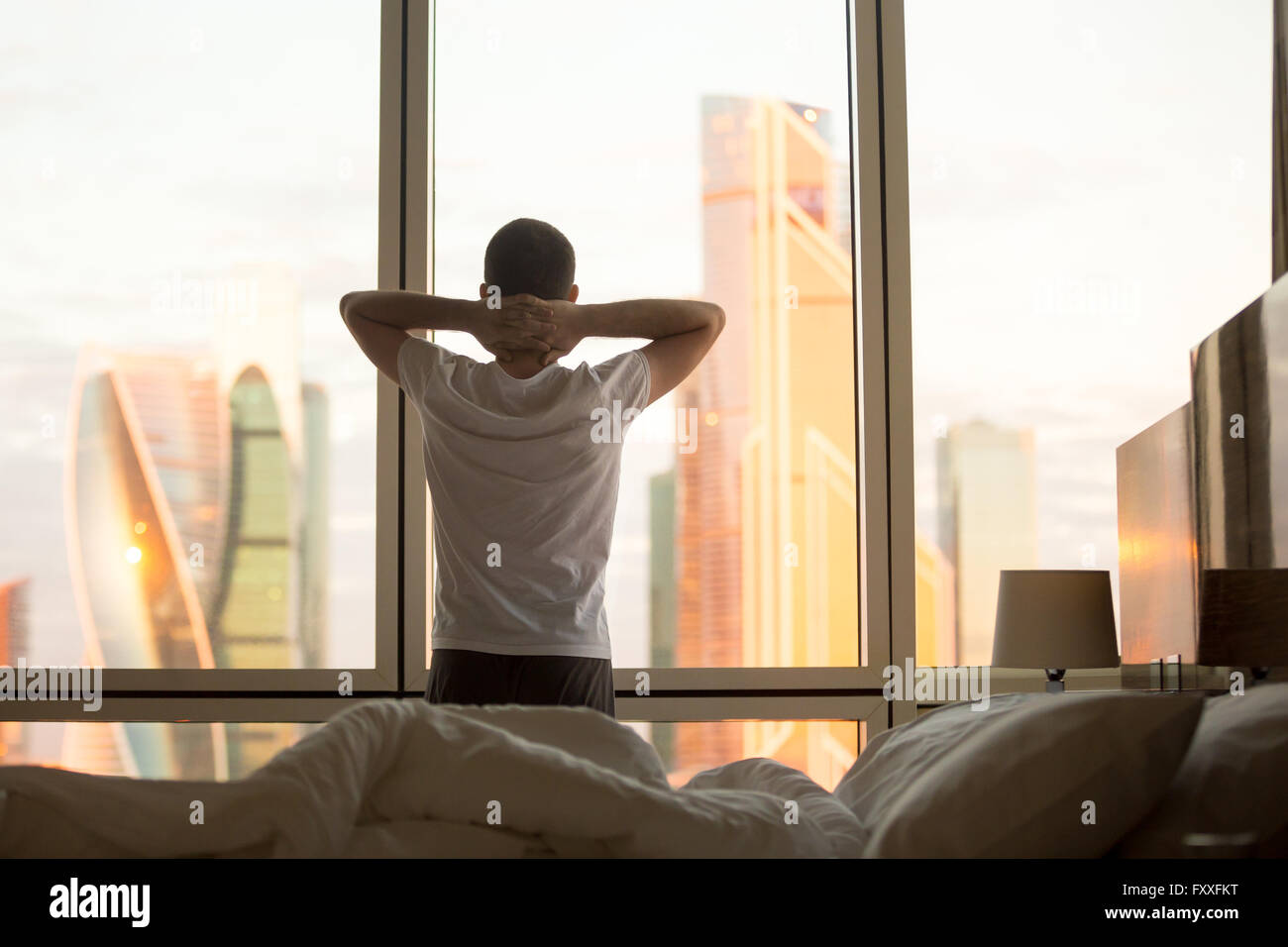 Rear view of young man looking at dawn city scenery in window after waking up. Handsome casual guy relaxing in the morning Stock Photo