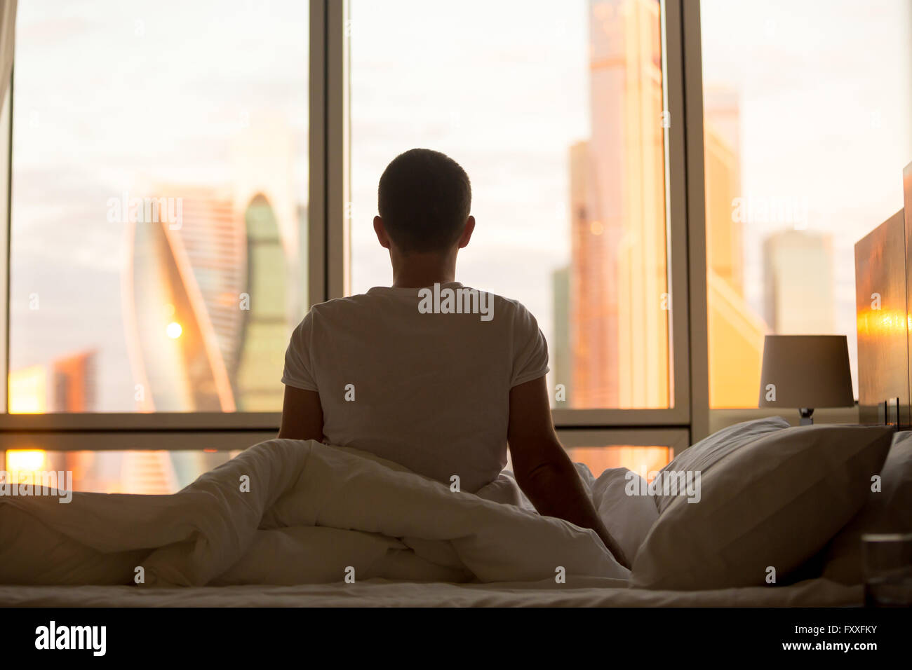 Rear view of young man sitting on unmade bed and looking at dawn city scenery in window after waking up. Handsome casual guy Stock Photo