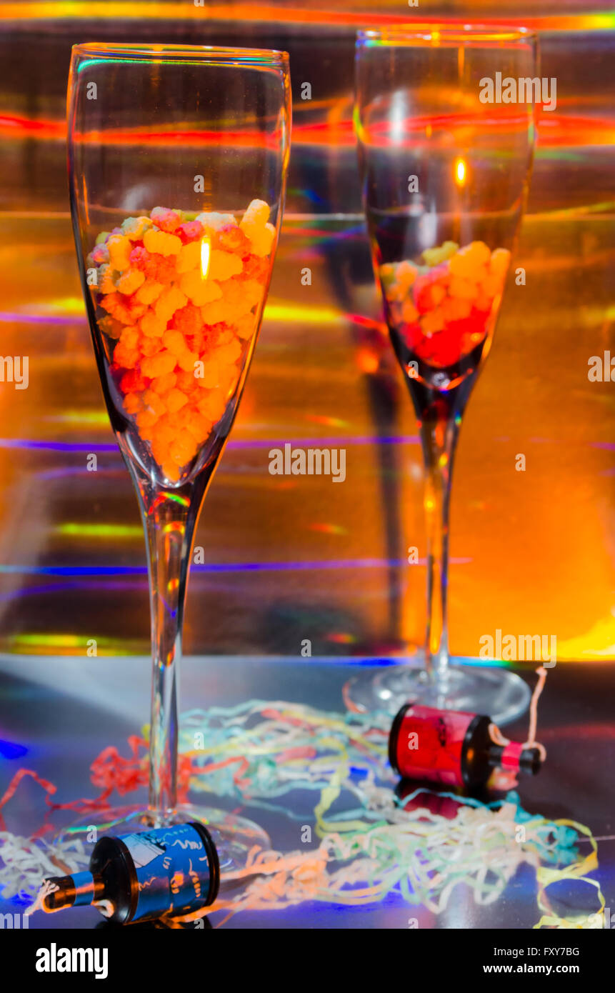 A Studio Photograph of Two Champagne Flutes Containing Sweets and Party Poppers Set Against an Orange Holographic Backdrop Stock Photo