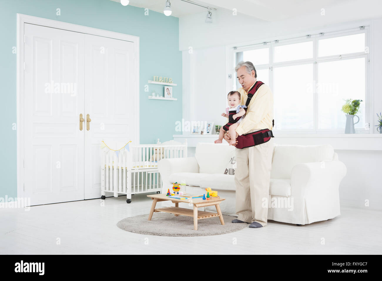 Senior man wearing a baby carrier and holding a baby in it to take care of the baby in a room Stock Photo