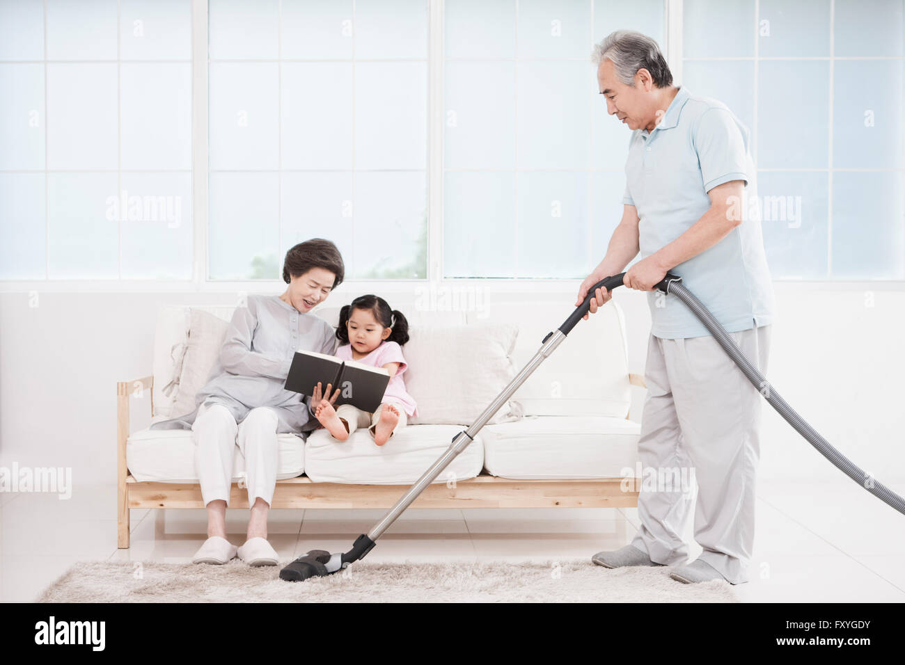Senior woman seated on a couch and reading a book with her granddaughter and her husband vacuuming on the floor Stock Photo