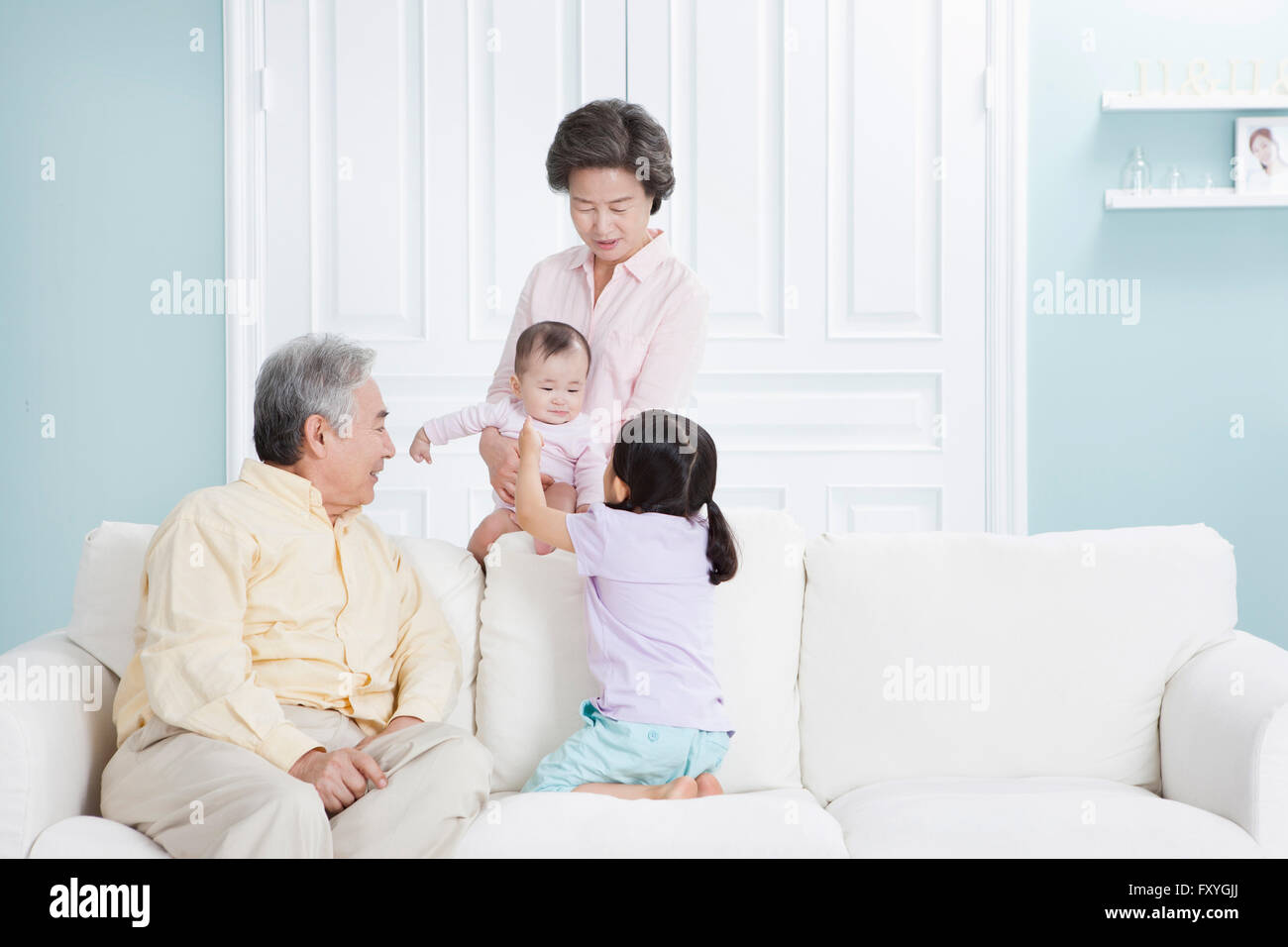 Grandmother holding a baby behind a couch and grandfather with granddaughter seated on a couch and looking at the baby with a smile Stock Photo