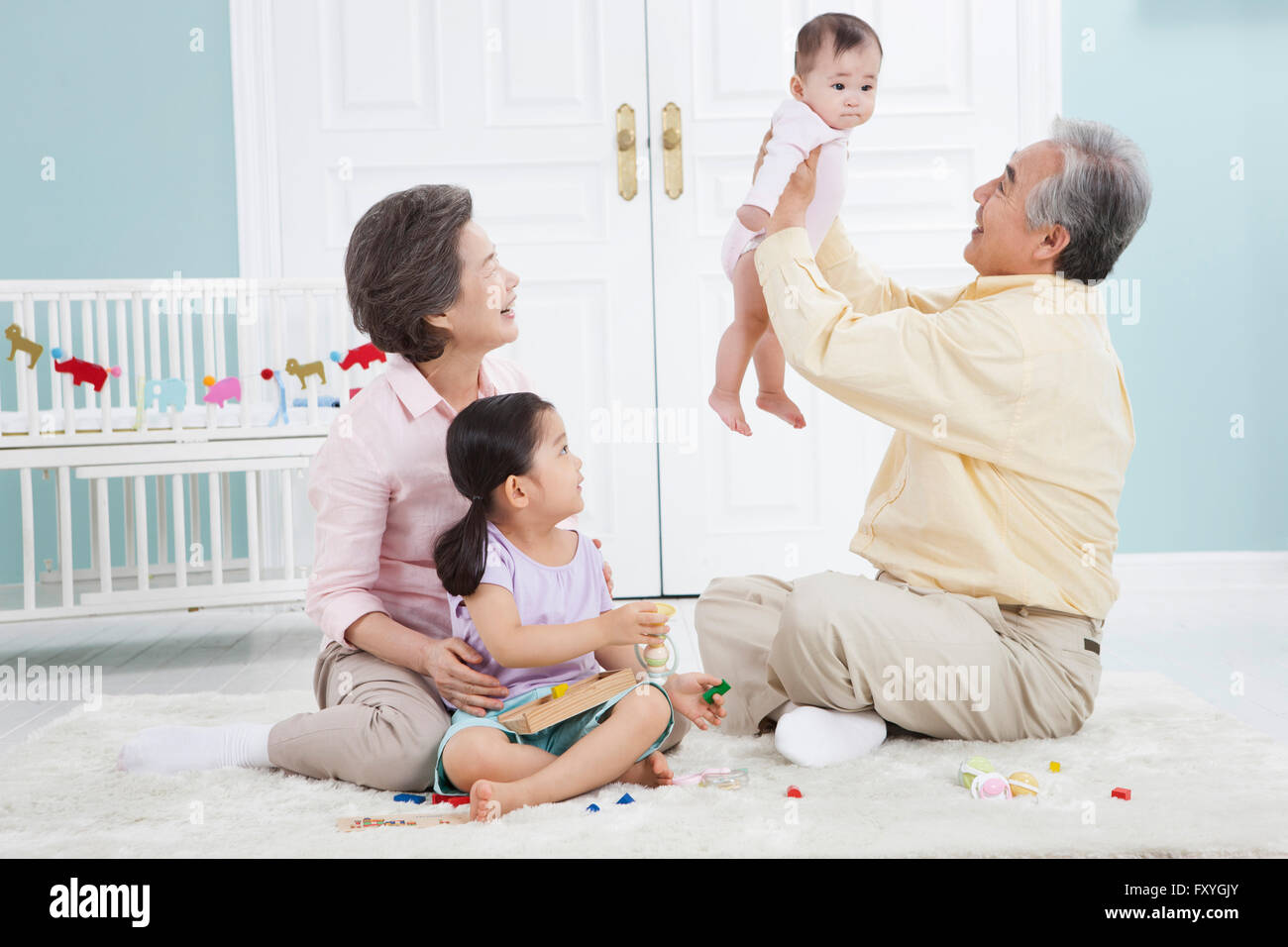 Grandfather holding a baby up and grandmother and granddaughter looking at the baby all seated on the floor Stock Photo
