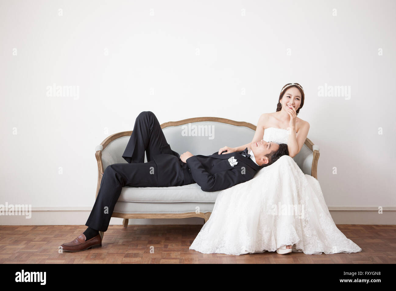 Groom lying on a couch with his head on bride's lap and bride smiling with her hand on her mouth Stock Photo