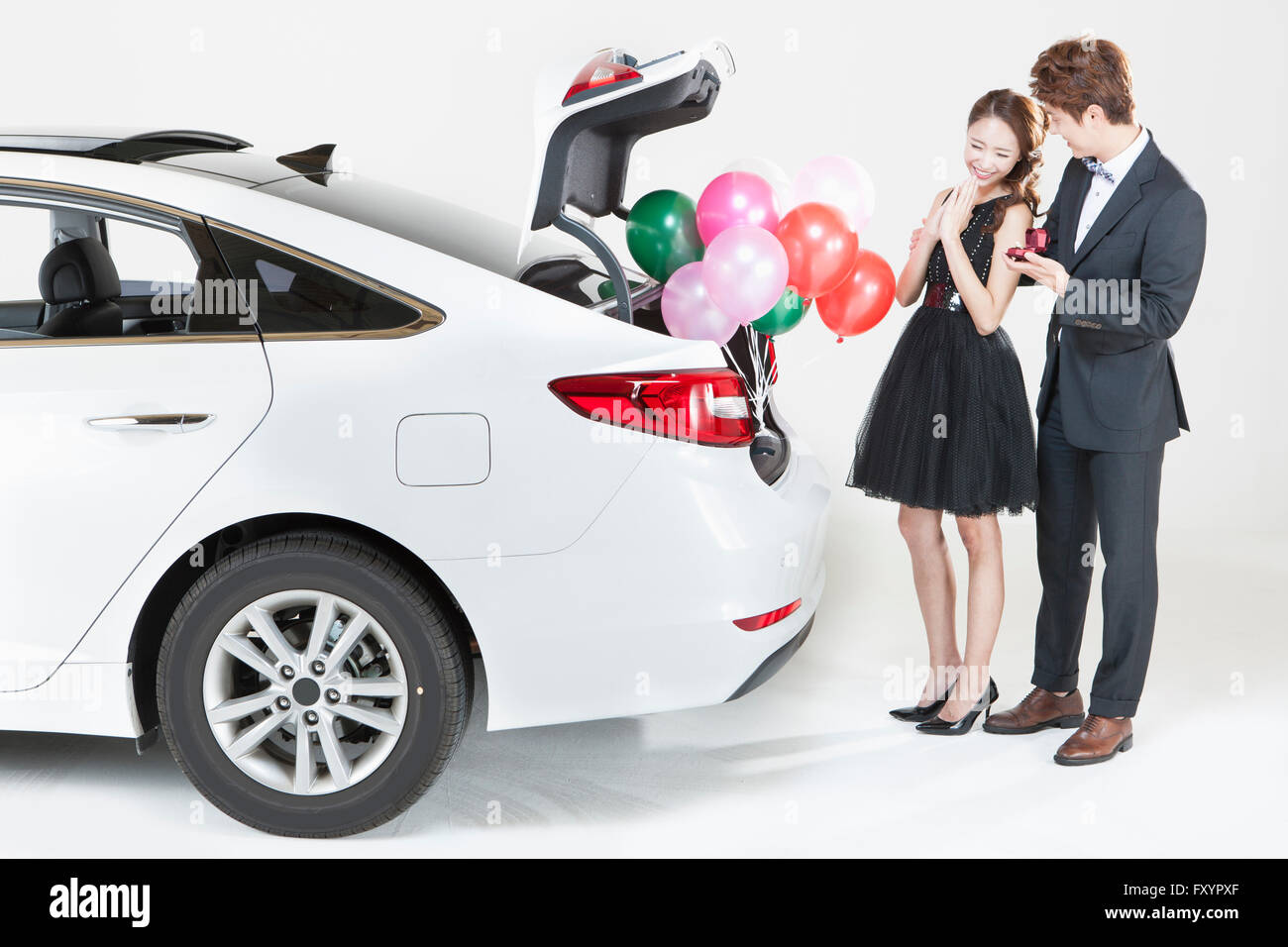 Woman delighted with present from man behind a catr with open trunk and balloons on it Stock Photo