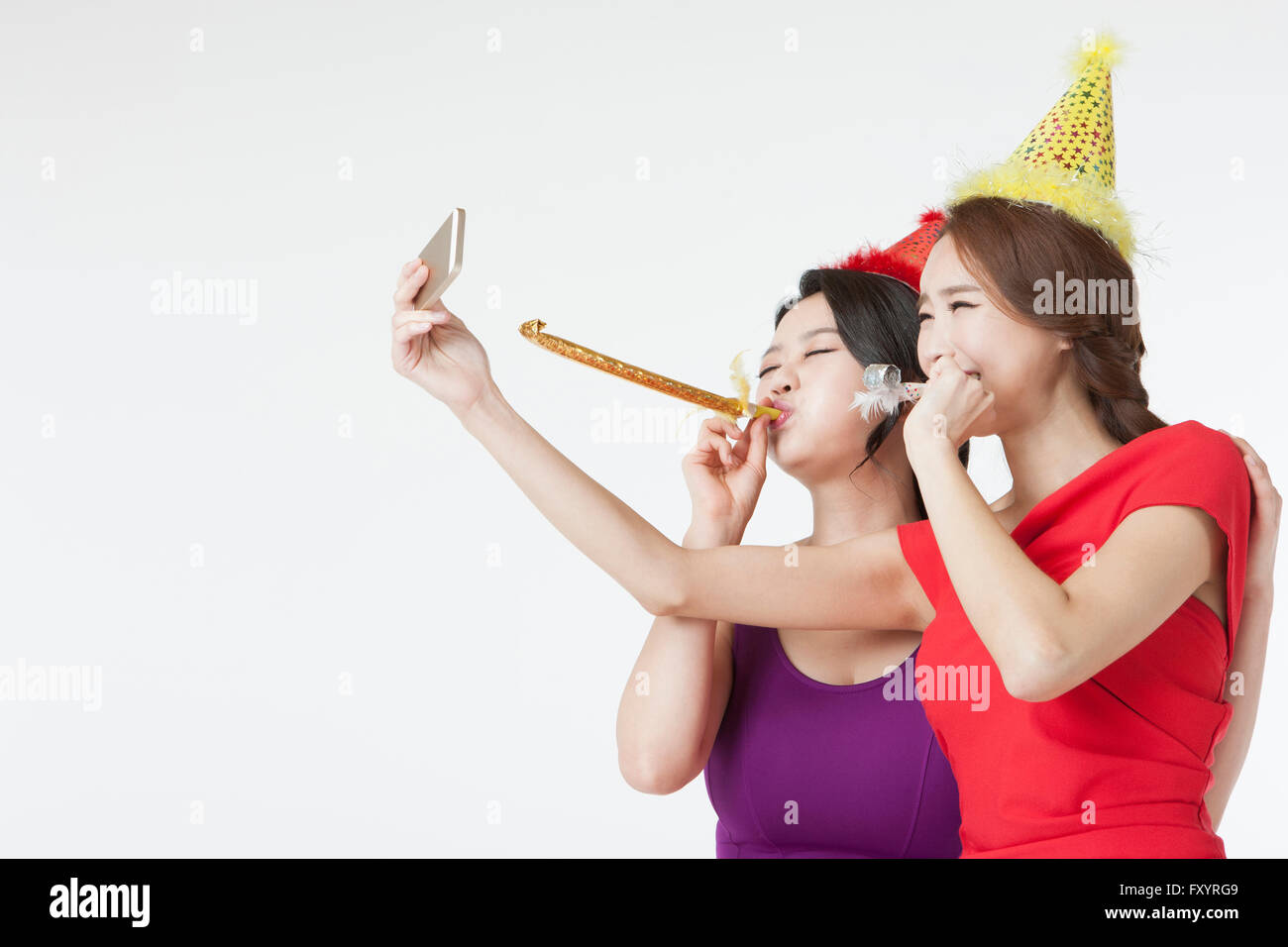 Side view portrait of two young smiling women talking a selfie blowing party horns Stock Photo