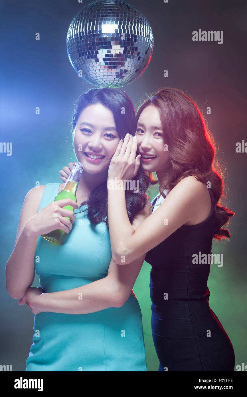 Two young smiling women talking in a whisper holding a beer bottle under a mirror ball staring at front Stock Photo