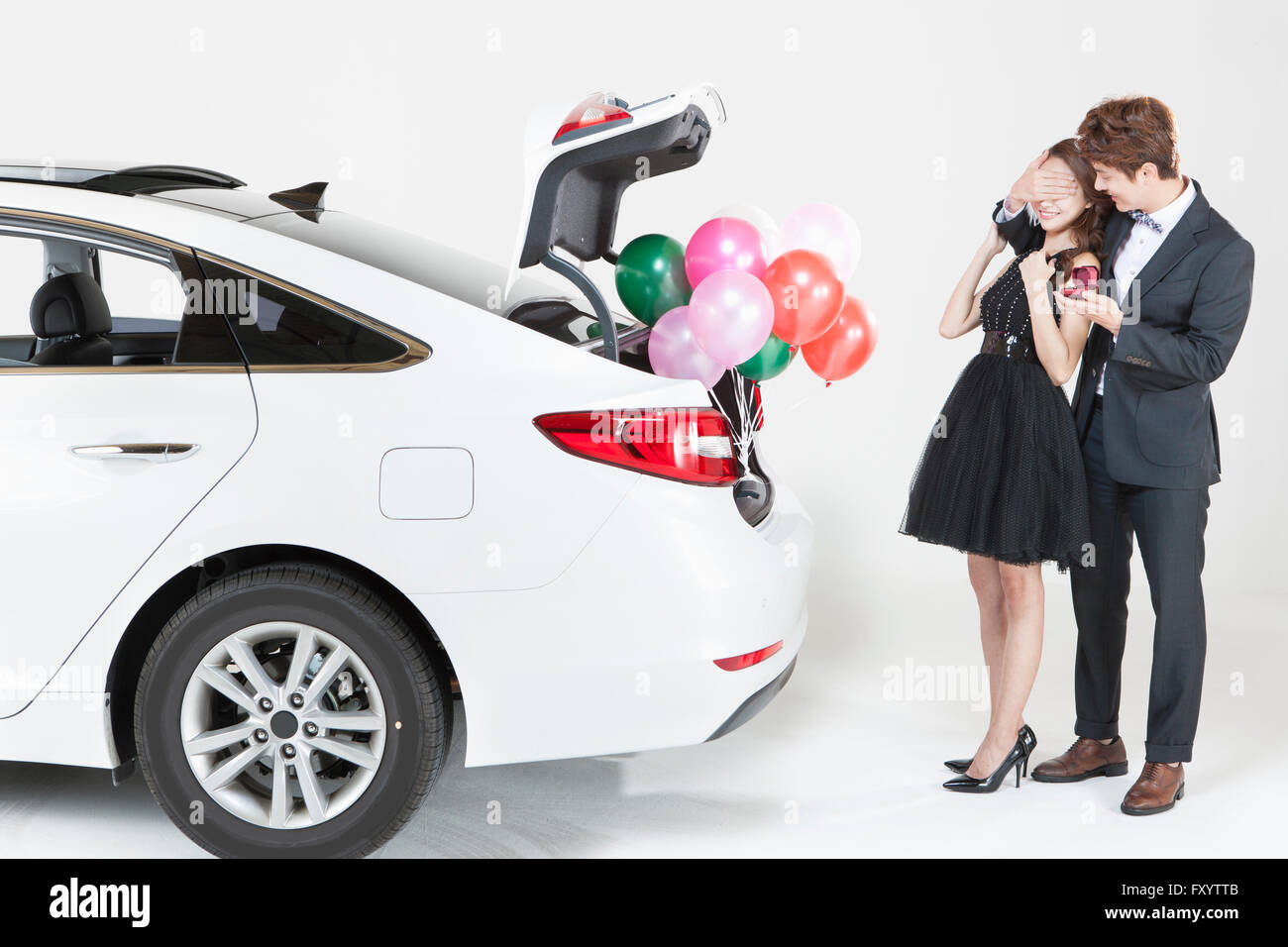 Young smiling man in suit holding a present box covering young woman's eyes in dress behind a car with balloons Stock Photo