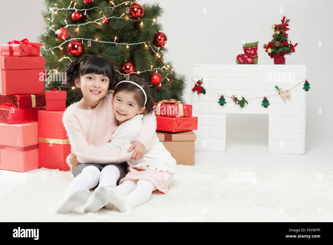Two smiling girls sitting down hugging each other with stacked present boxes, Christmas tree and fireplace Stock Photo