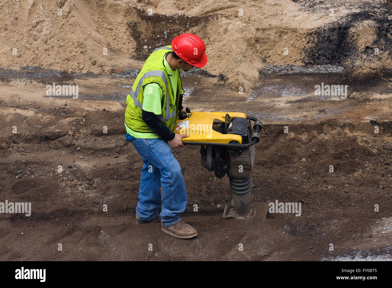 A Construction Worker operating a Wacker Neuson BS60-4S gas powered rammer tamping machine inside of a ditch dug into the street Stock Photo