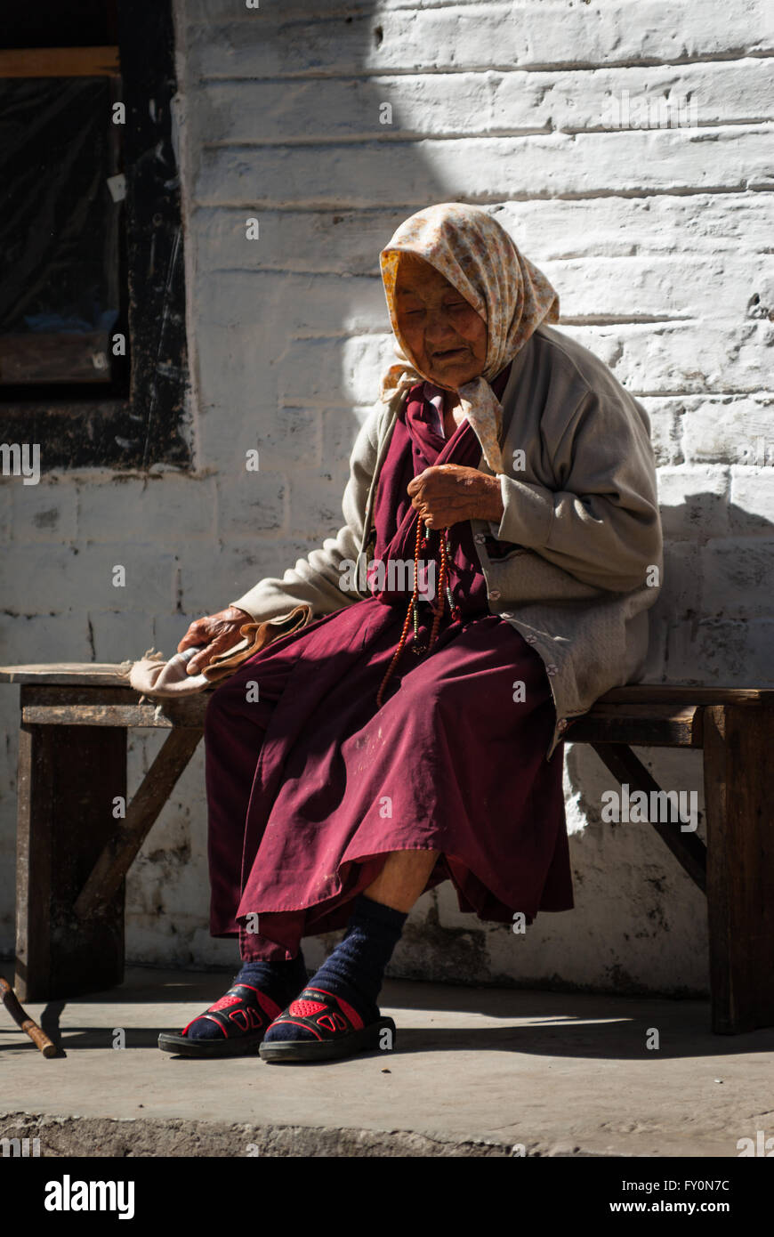 Elderly woman wearing crimson robe and holding Buddhist prayer beads sitting on wooden bench in Trongsa, Bhutan Stock Photo