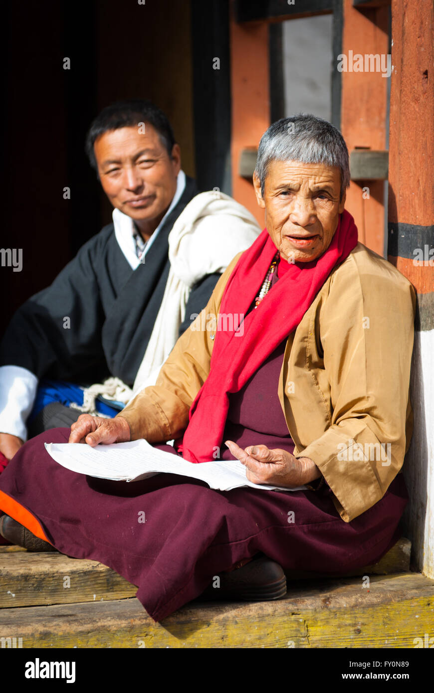 Elderly Vajrayana Buddhist nun in crimson robe and Bhutanese man in traditional gho sitting on terrace at Paro Dzong, Bhutan Stock Photo