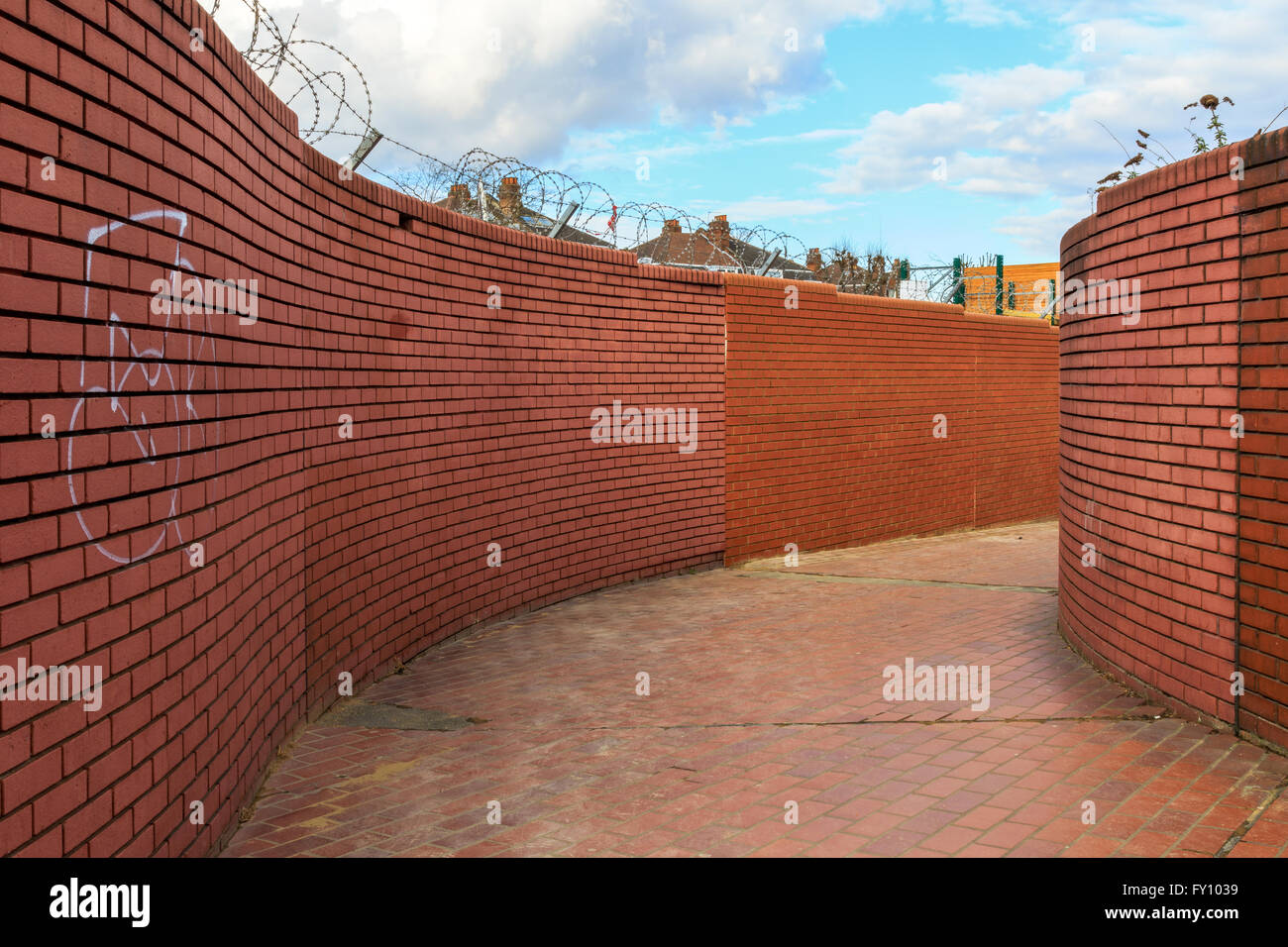 Curving walled path with graffitied red brick walls topped with barbed wire Stock Photo