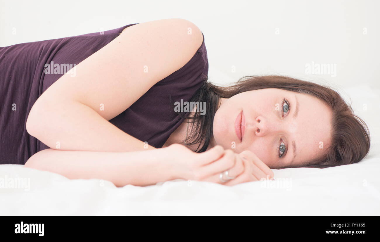Portrait of young woman lying down in her bed resting. She is looking at camera with a serious and bored expression. Stock Photo