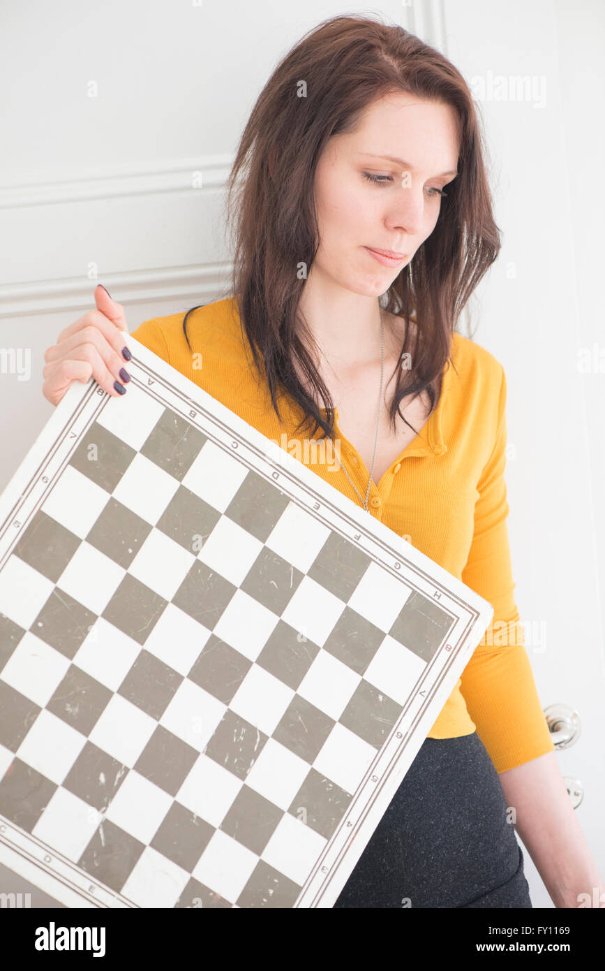 Serious woman holding chessboard in home interior. Concept of strategy, leisure activity and contemplation. Stock Photo