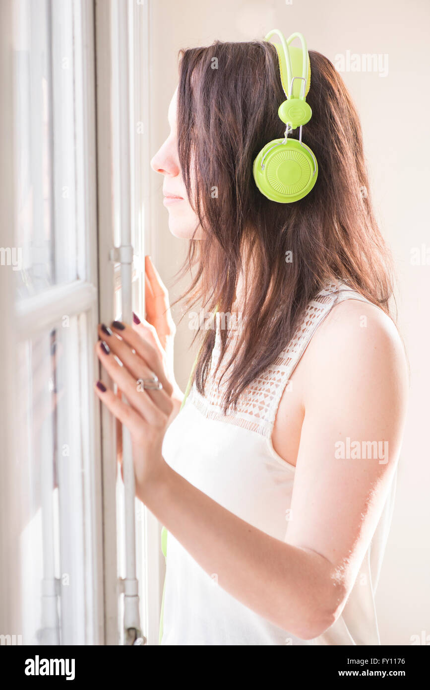 Woman standing by a window, looking away. She is listening to music in green headphones. Lifestyle image of carefree relaxation. Stock Photo