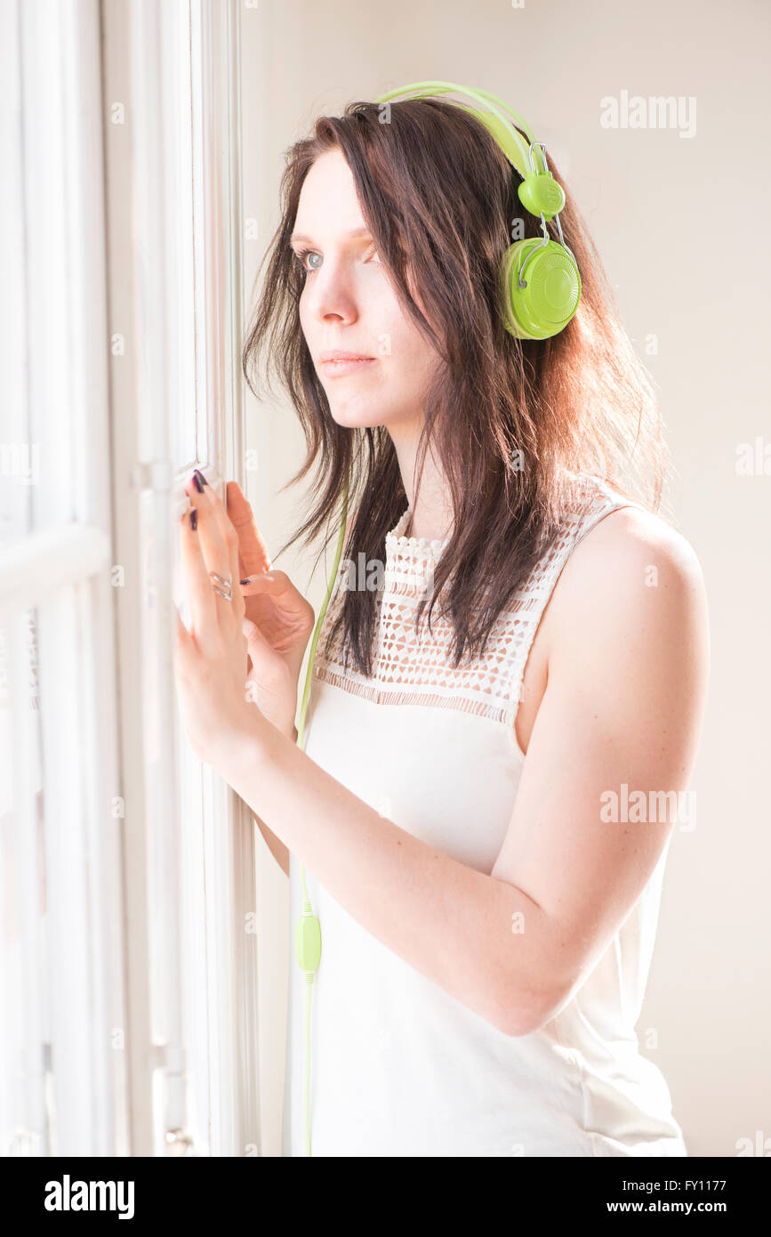 Woman standing by a window, looking away. She is listening to music in green headphones. Lifestyle image of contemplation. Stock Photo