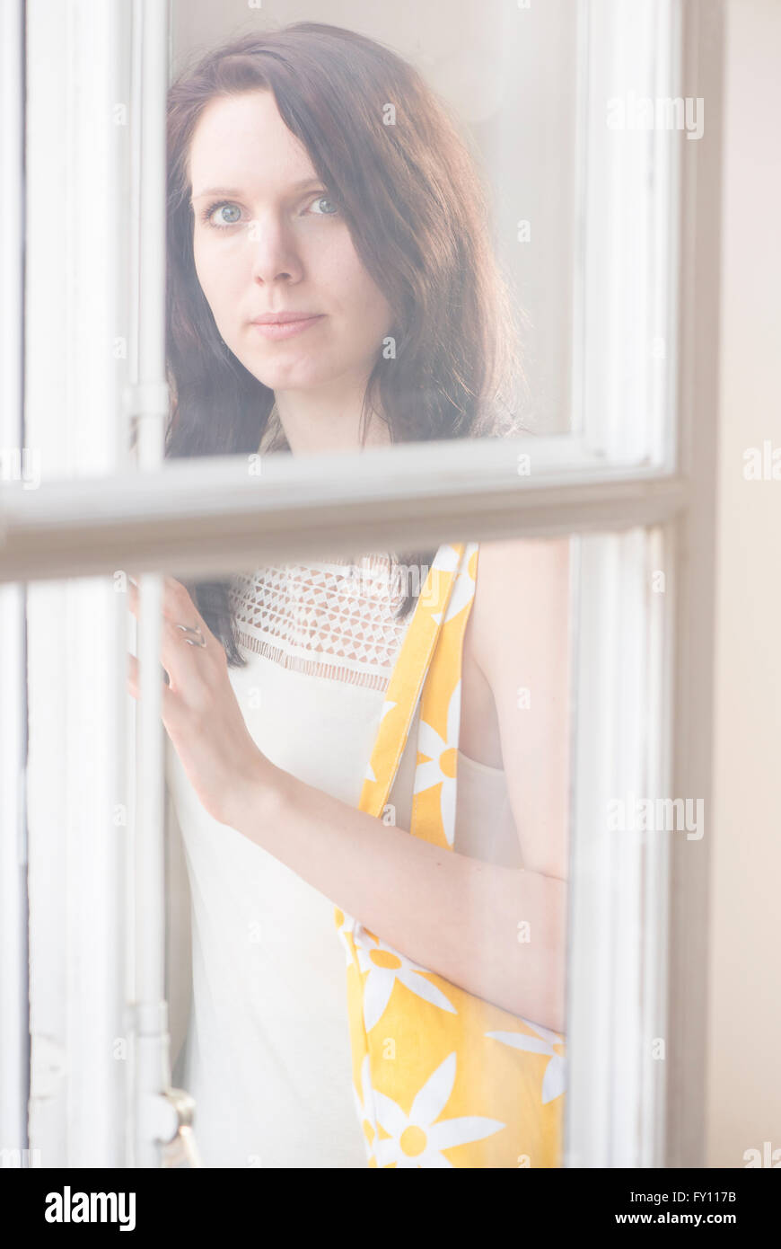 Woman standing by a window, looking at camera. Concept of sadness, waiting and anticipation. Lifestyle image of contemplation. Stock Photo