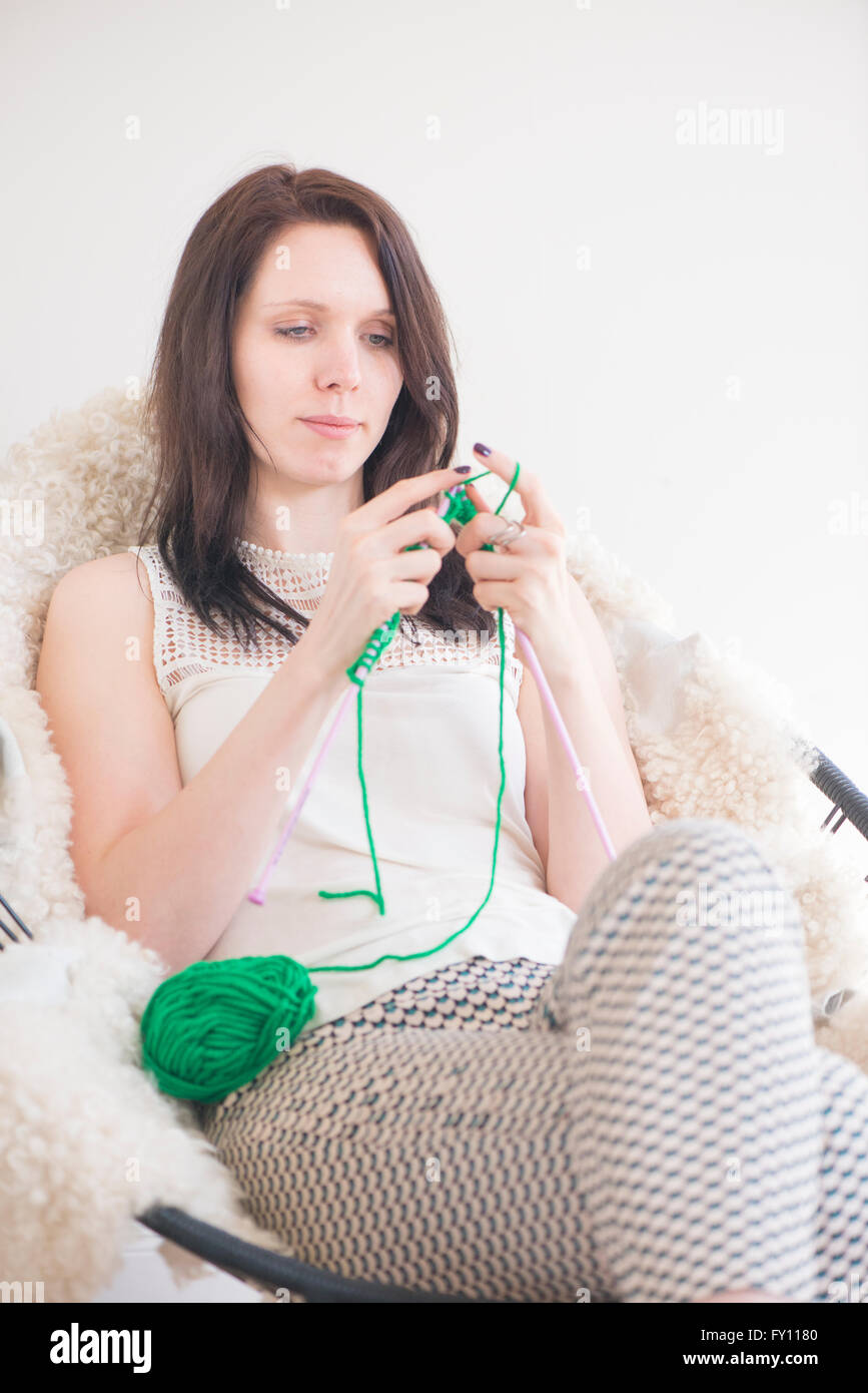 Woman knitting. She is sitting in a chair in home interior. Lifestyle image showing leisure activity and hobby. Stock Photo