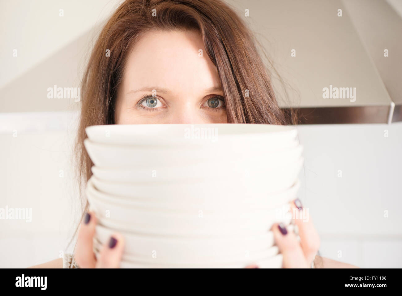 Portrait of woman in kitchen holding a stack of plates. Lifestyle image of young woman. Concept of dinner or party preparation. Stock Photo