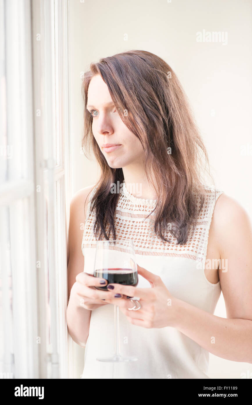 Woman standing by a window, looking away. Concept of sadness, waiting and anticipation. Lifestyle image of contemplation. Stock Photo