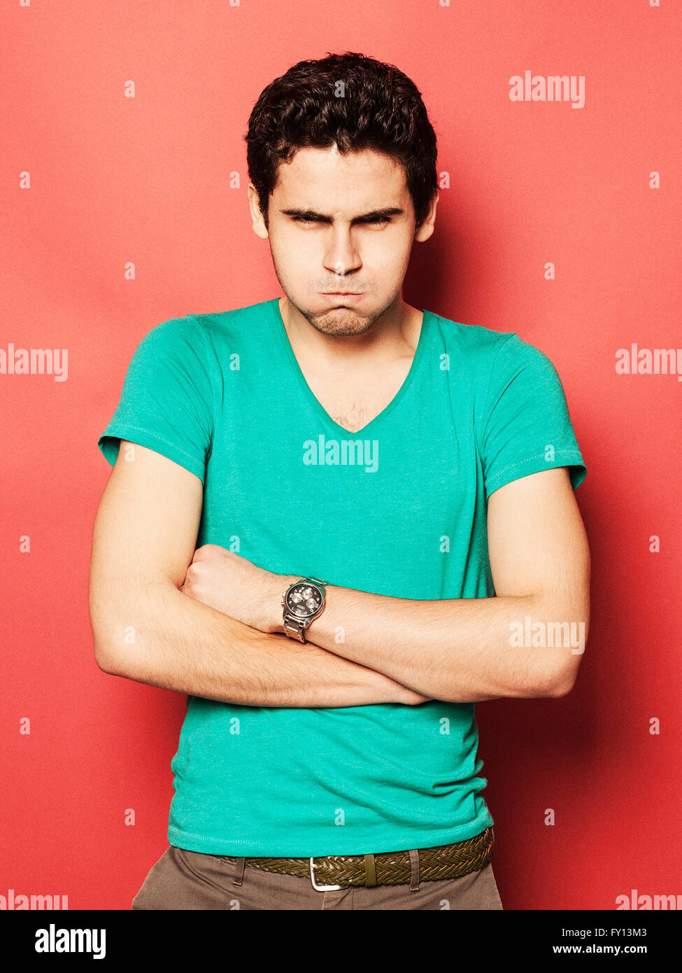 Portrait of angry young man with puffed cheeks standing arms crossed against red background Stock Photo