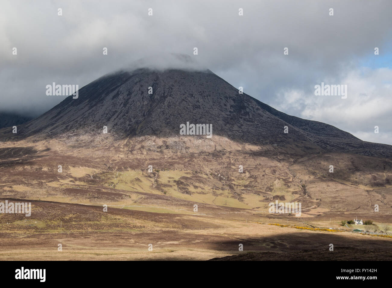Barren Scottish mountain with the top covered in clouds. Small white house at the bottom. Stock Photo